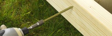 Close-up of a green hex head timber screw being driven into a wooden plank using a power drill, on a grassy outdoor surface