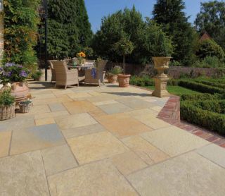 Sunlit garden patio paved with Pavestone calibrated limestone slabs in a blend of warm golden, cream, and light grey tones. The patio features rattan garden furniture, terracotta planters, and overlooks a manicured lawn bordered by box hedging and mature trees