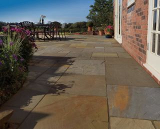 Charming garden patio featuring Pavestone Calibrated Sandstone in Golden Fossil, with a rich mix of buff, ochre, and slate-grey tones. The paving leads to a dining area with wrought iron furniture, surrounded by vibrant flower borders and terracotta pots, set beside a traditional red brick home with countryside views