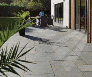 Modern garden patio paved with Pavestone Kota Blue limestone slabs in a uniform layout, adjoining a contemporary home with large glass doors. The space features black rattan dining furniture, potted palm plants, and a neatly trimmed lawn in the background
