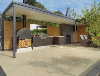 Modern outdoor kitchen and dining area featuring Pavestone Classic Porcelain Paving Tiles in a light beige tone. The clean-lined porcelain tiles provide a sleek, durable surface beneath a covered BBQ zone with a wood-fired oven and contemporary rattan seating. Surrounded by timber cladding and landscaped greenery, perfect for stylish garden entertaining