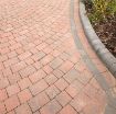Close-up view of Brett Alpha 50mm block paving in Brindle, arranged in a curved pattern with a contrasting charcoal border. The paving leads to a raised flower bed with blooming plants and bark mulch, highlighting a neat and decorative driveway or garden edge