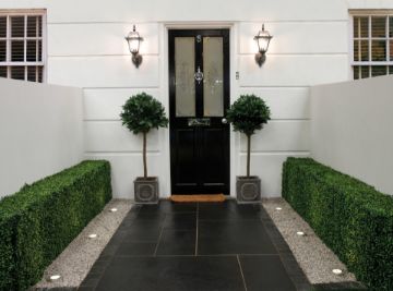 Elegant entranceway featuring Pavestone Black Kadapha limestone paving laid in a clean linear pattern, bordered by gravel strips with recessed uplighting and neatly clipped hedges. Two topiary trees in square planters frame a black front door with traditional lantern wall lights