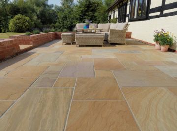 Spacious countryside patio laid with Pavestone Calibrated Sandstone in Rustic Blend, showcasing warm tones of buff, amber, and soft plum. The patio features a wicker outdoor seating set, red brick retaining walls, and potted flowers, all set against a traditional timber-frame cottage