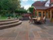 Elegant garden patio featuring Pavestone Calibrated Sandstone in Modak, displaying warm tones of terracotta, rose, and muted plum. The outdoor space includes curved stone steps, a wooden dining set, and landscaped borders, with a spaniel resting on the raised lawn beside a vintage bistro set
