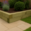 Raised garden bed constructed from pressure-treated timber sleepers stacked neatly on a sandstone patio, surrounded by grass and planted with lavender, boxwood, and leafy shrubs in a landscaped setting