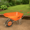 Bright orange Belle Warrior wheelbarrow positioned on a compacted earth surface in a well-maintained garden, with lush grass, green shrubs, and a timber border in the background