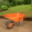 Bright orange Belle Warrior wheelbarrow positioned on a compacted earth surface in a well-maintained garden, with lush grass, green shrubs, and a timber border in the background